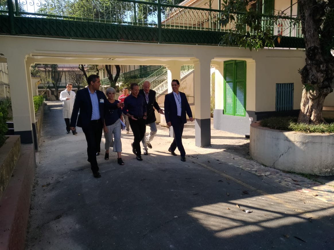 Imagem colorida, com sete pessoas no meio está o Secretário da Saúde Edson Aparecido, ao seu lado direito o Subprefeito de Jaçanã/Tremembé, Alexandre Pires, estão passando por baixo de uma passarela no Hospital Geriátrico Dom Pedro II.